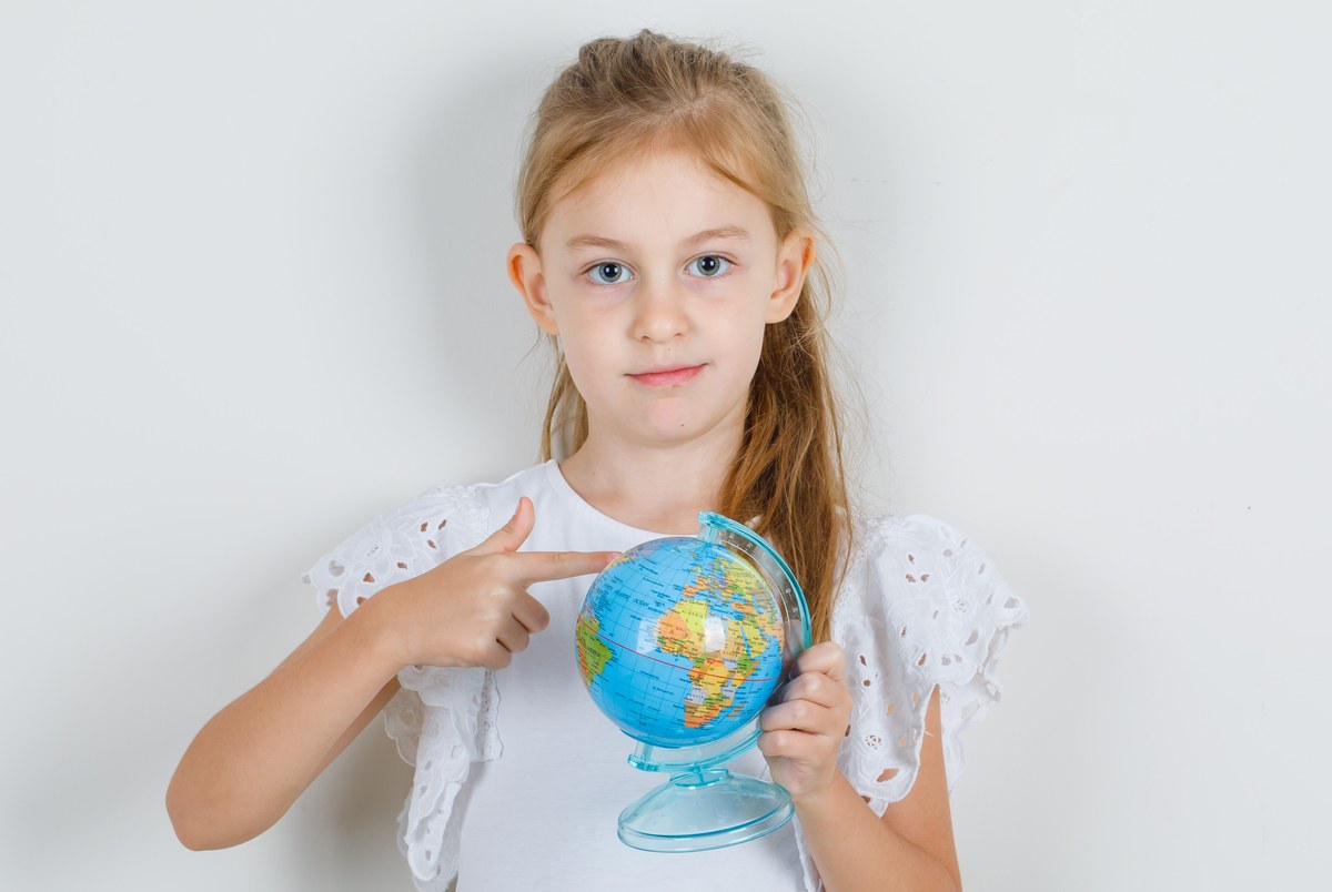 Little girl showing globe with finger in white t-shirt, skirt , front view.